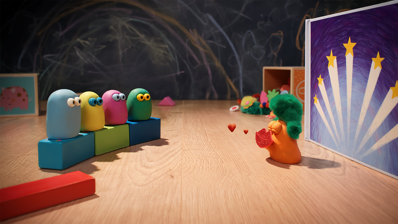 Blue, Yellow, Pink and Green are sitting on building blocks facing a background for a stage. Orange is in front wearing a green wig and glitter red lips 